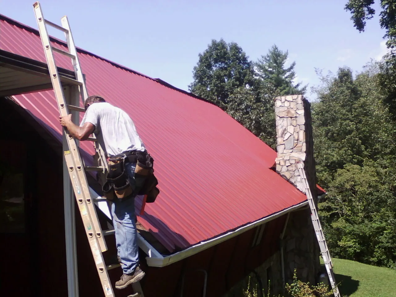 Roofer on a ladder beside a red metal roof and stone chimney — Soffit & Fascia Repair by Roofing Brothers in Moab